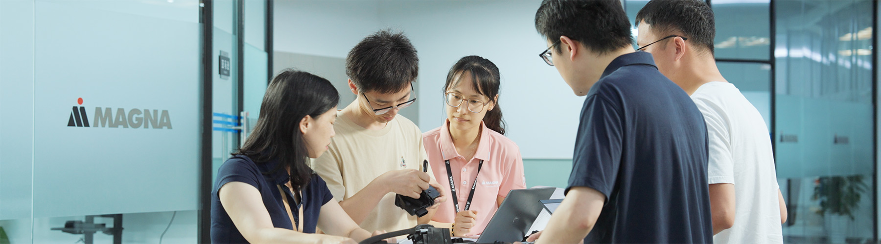 Five people standing around a table with a lap top and examining an automotive part in an office environment