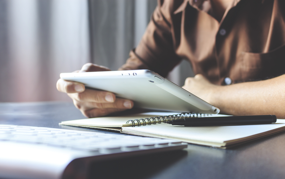 Person using a tablet at a desk with a notebook, pen, and keyboard