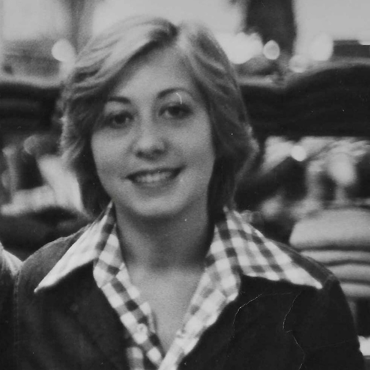 Smiling woman with short hair in a black-and-white photo, wearing a checkered shirt and jacket. The background is blurred, conveying a warm, nostalgic tone.