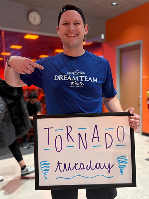Luke indoors pointing at a Tornado Tuesday workout sign while wearing a Make-A-Wish Dream Team shirt