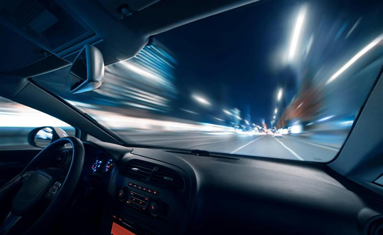 View from inside a car driving fast at night. Blurred lights and streaks outside the windshield convey speed and motion. Dashboard is dimly lit with driver monitoring technology in the interior mirror.